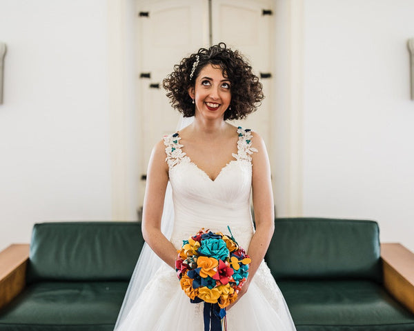 Cathy from The Origami Boutique on her wedding day as a smiling bride in a white wedding dress stands indoors holding a vibrant multicolour paper flower bouquet. She is positioned in front of a dark green couch and white walls.