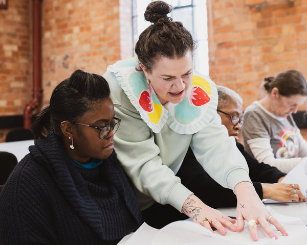 Two women working together at a table in a room with brick walls and a sign that reads 'Watling'.